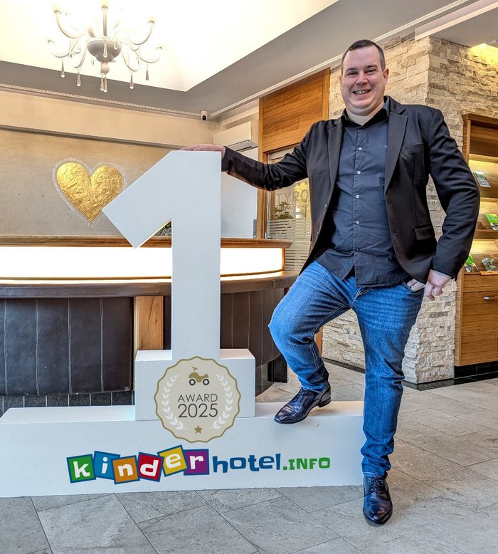 TYROL Family Retreat. Mehr als ein Familienhotel in Naturns. Mann steht an der Rezeption neben einem großen 1st-Place-Schild für kinderhotel.info Award 2025