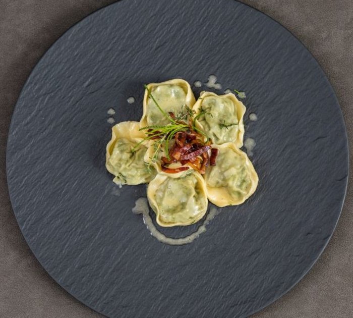 Pure Flexibilität im TYROL Family Retreat Fünf gefüllte Ravioli auf schwarzem Teller mit Kräutern und Speck als Garnitur