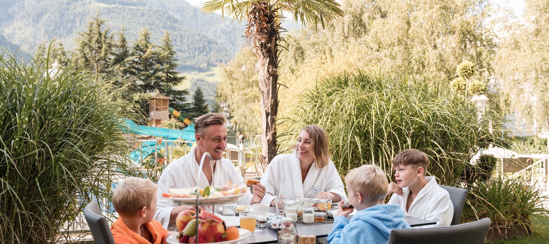 Pure Flexibilität im TYROL Family Retreat Familie frühstückt im Außenbereich mit Bergen und Palmen im Hintergrund
