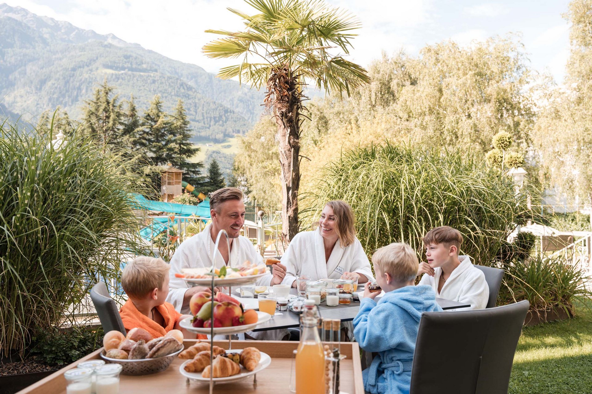 TYROL Family Retreat als Familienhotels in Südtirol Familie frühstückt im Außenbereich mit Bergen und Palmen im Hintergrund