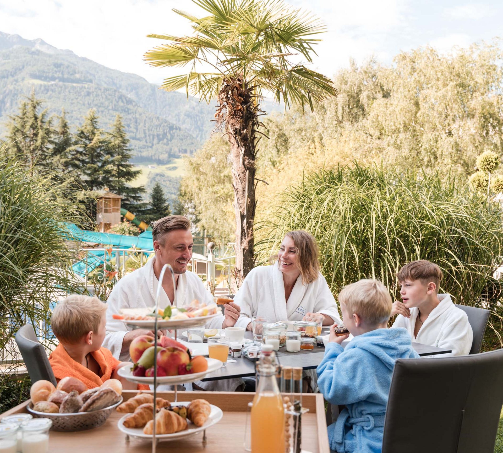 Königlicher Urlaub im Aparthotel nahe Meran und Umgebung Familie frühstückt im Außenbereich mit Bergen und Palmen im Hintergrund