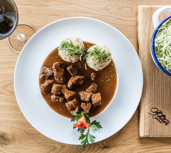 Pure Flexibilität im TYROL Family Retreat Gulasch mit zwei Semmelknödeln, Beilagensalat und Rotwein auf Holztisch