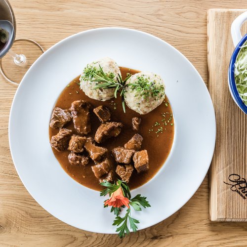 Pure Flexibilität im TYROL Family Retreat Gulasch mit zwei Semmelknödeln, Beilagensalat und Rotwein auf Holztisch