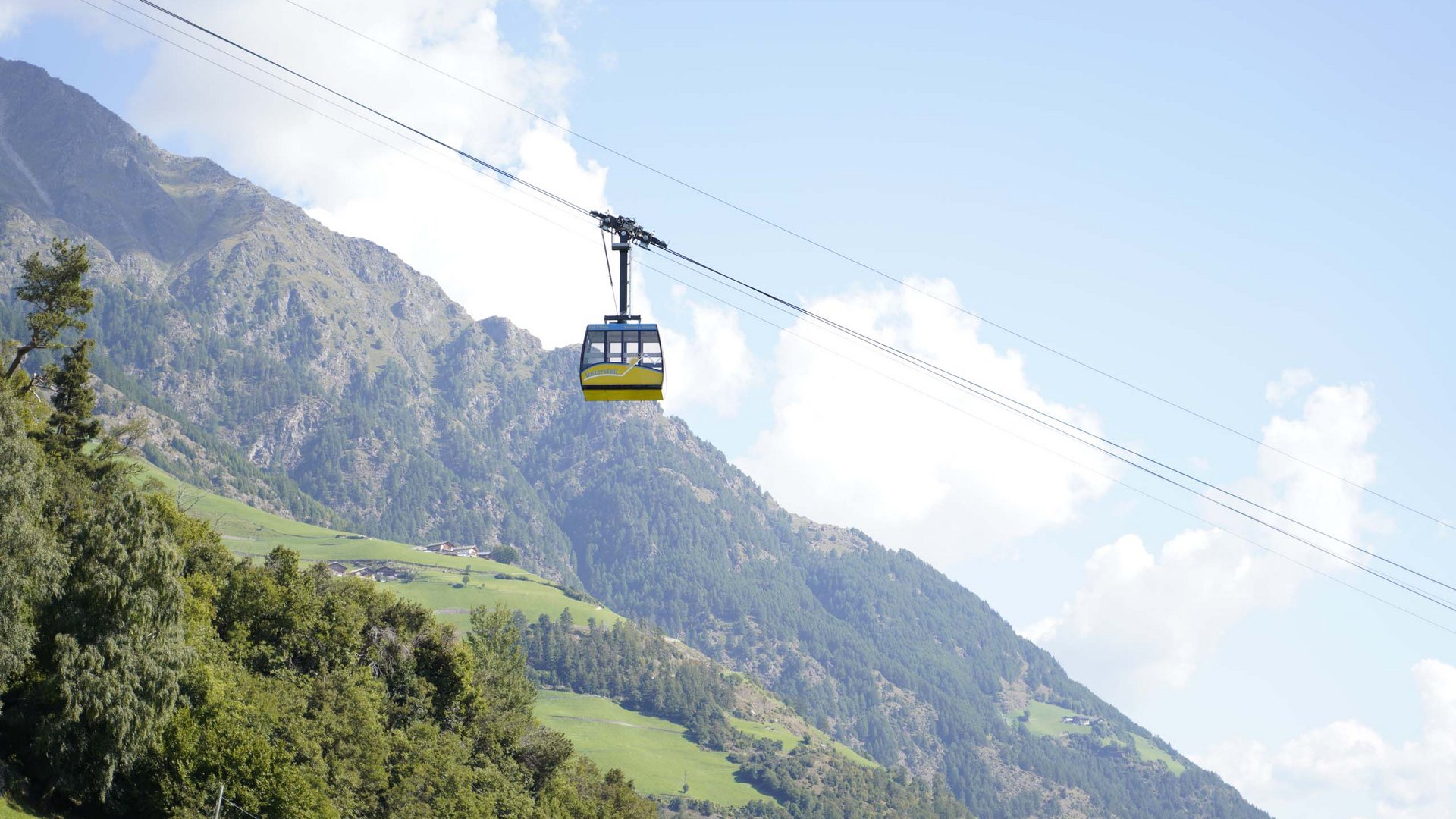 Euer vielseitiger Urlaub in Naturns Seilbahn fährt über grüne Alpenlandschaft mit Bergen und blauem Himmel