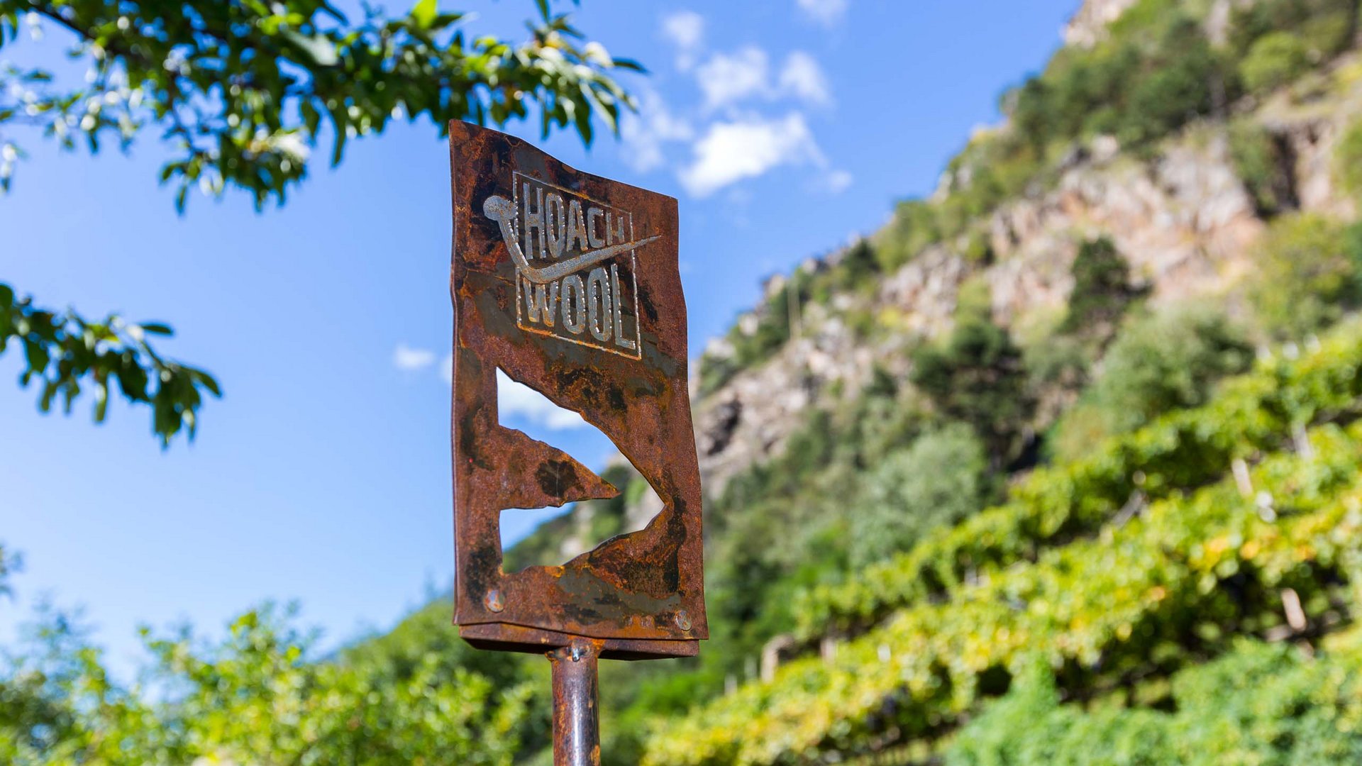 Euer vielseitiger Urlaub in Naturns Verrostetes Metallschild mit Text vor grün bewachsenem Hügel und blauem Himmel