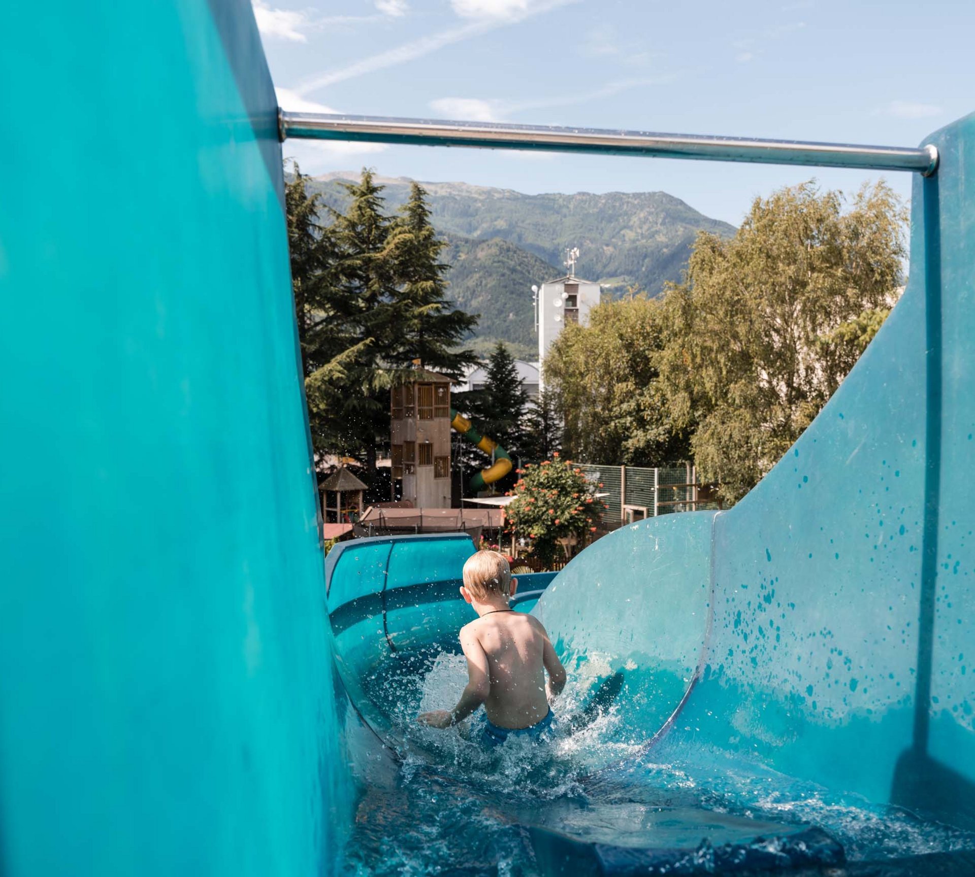 Ab ins Familienhotel in Südtirol mit Wasserrutsche! Kind rutscht auf einer blauen Wasserrutsche im Freibad mit Bergblick