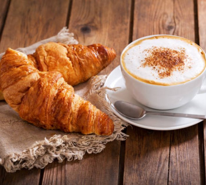Pure Flexibilität im TYROL Family Retreat Zwei Croissants auf einem Tuch neben einer Tasse Cappuccino auf Holztisch