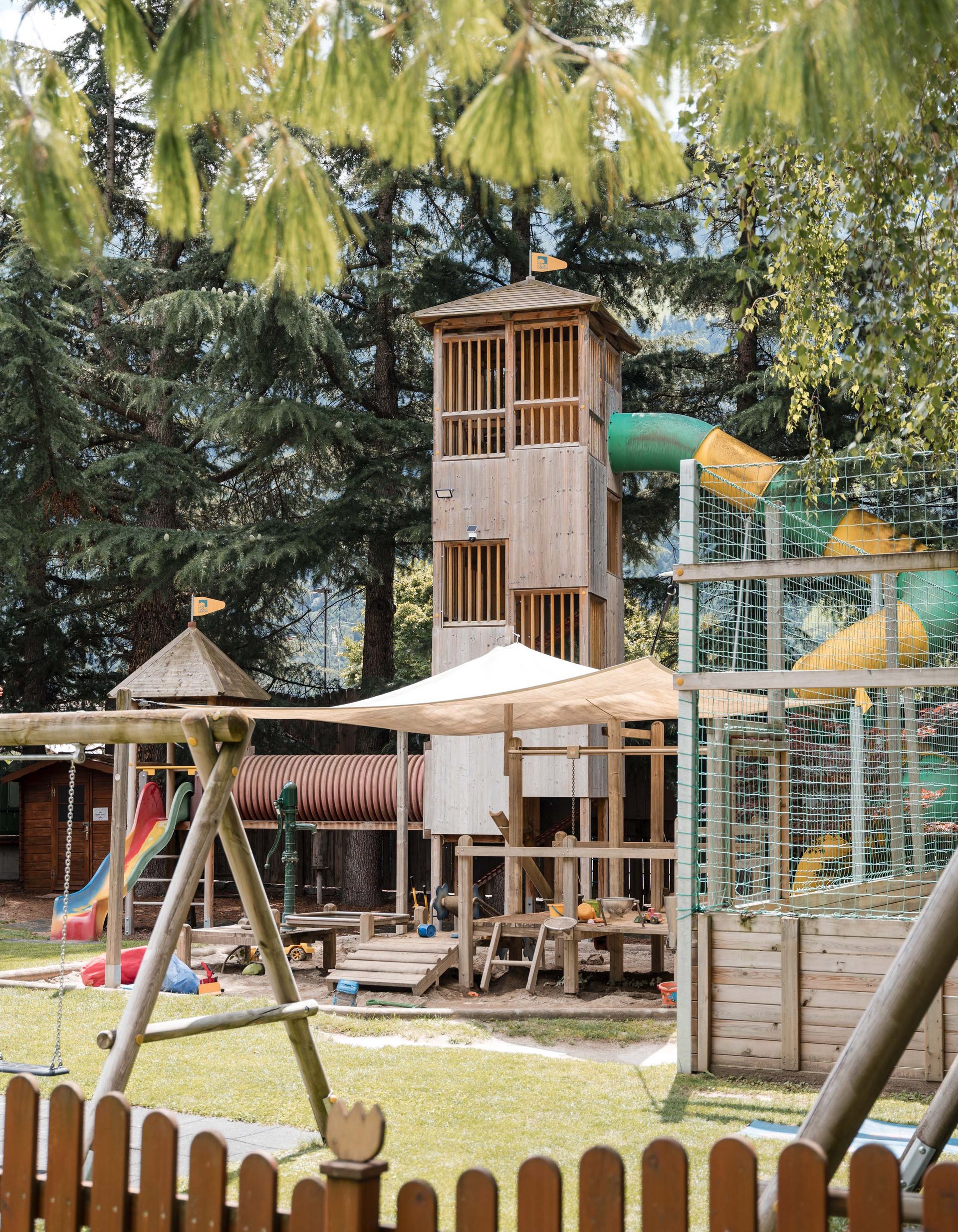 TYROL Family Retreat. Mehr als ein Familienhotel in Naturns. Kinderspielplatz mit Holzspielturm, Rutsche und Schaukel umgeben von Bäumen