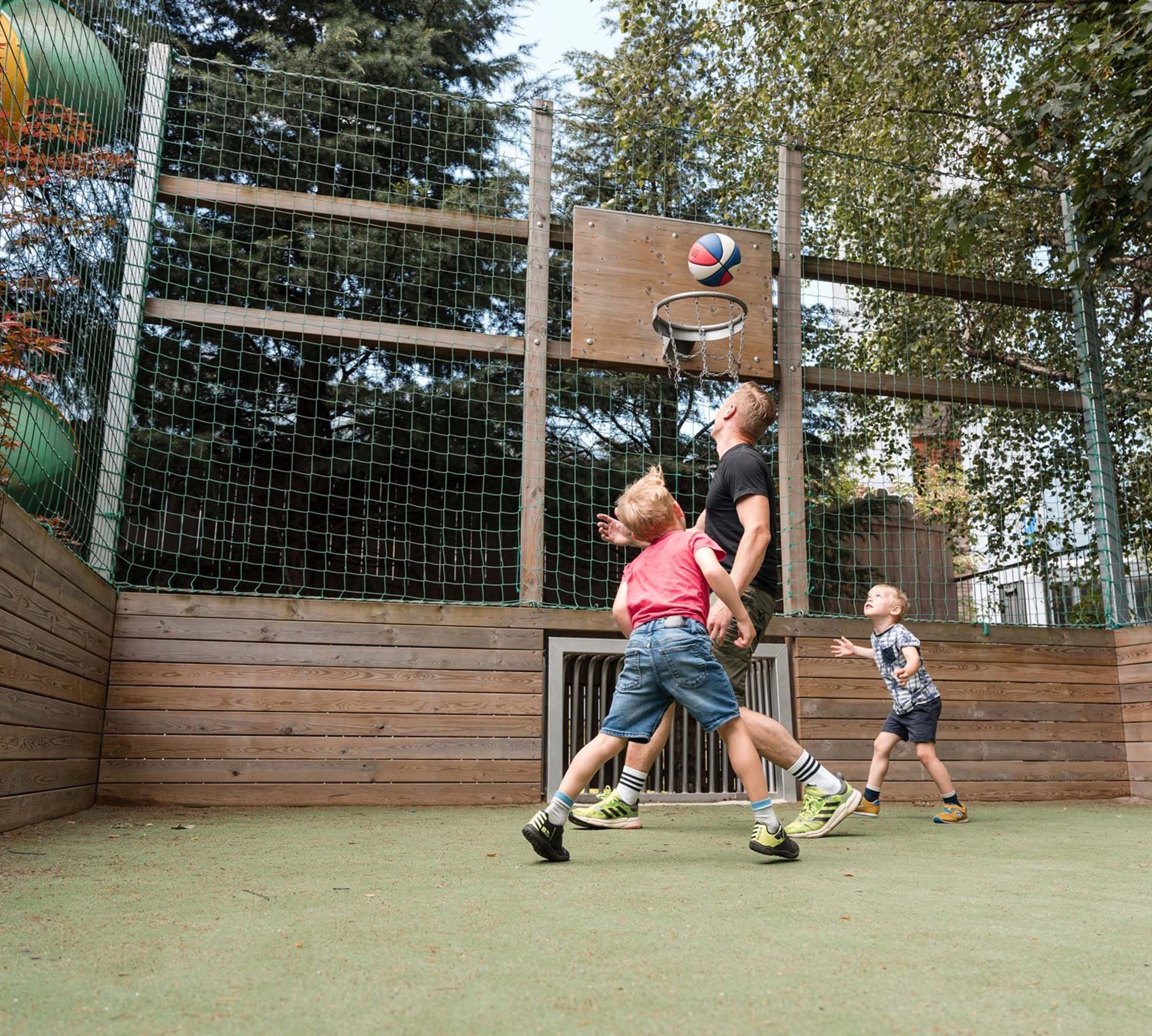Spiel und Spaß im Hotel mit Kinderbetreuung in Südtirol Vater und zwei Kinder spielen Basketball im Außenbereich