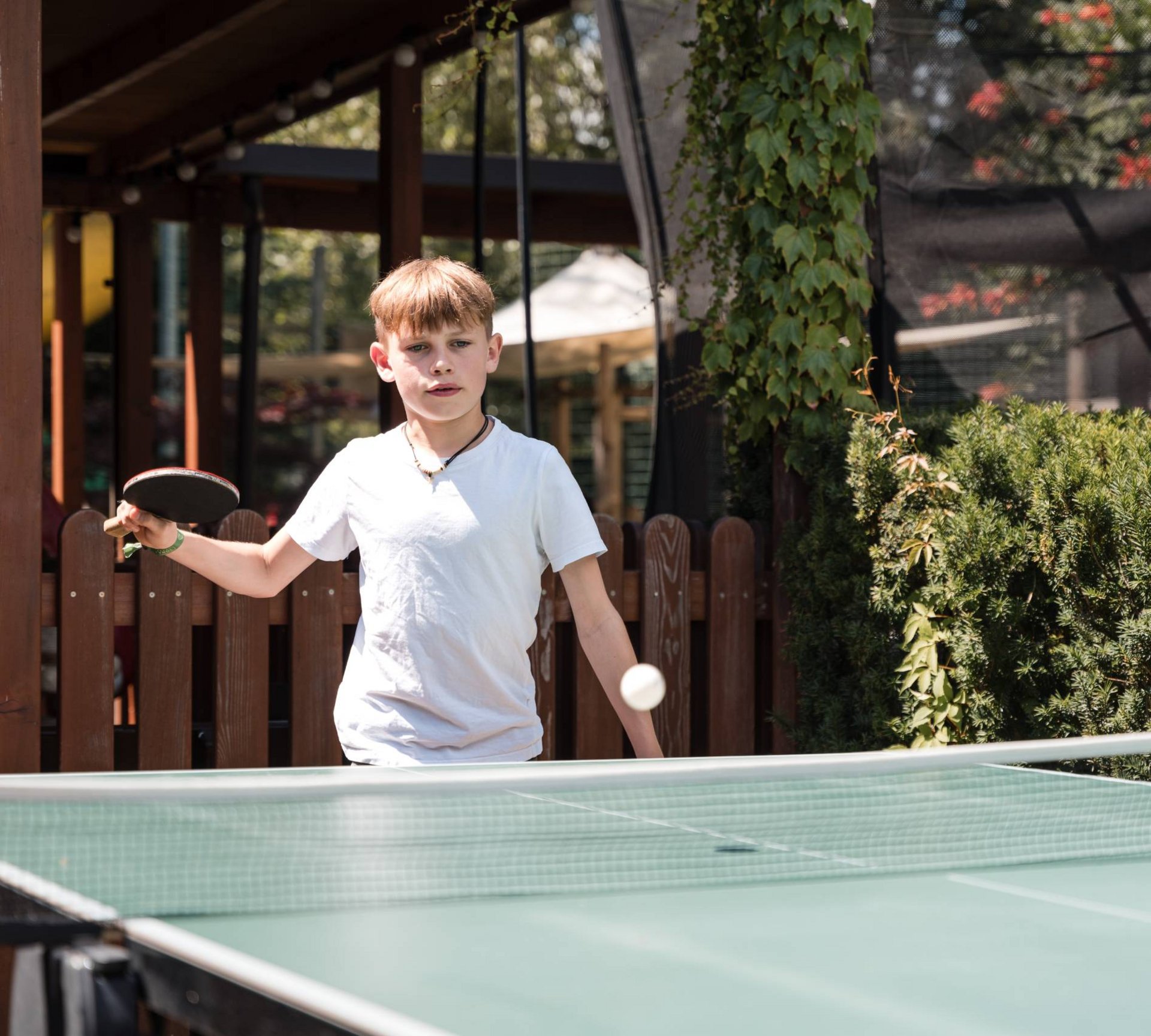 Abenteuer im Teenager-Hotel in Südtirol Junge spielt Tischtennis im Freien mit grünem Tisch und Umgebungsbepflanzung