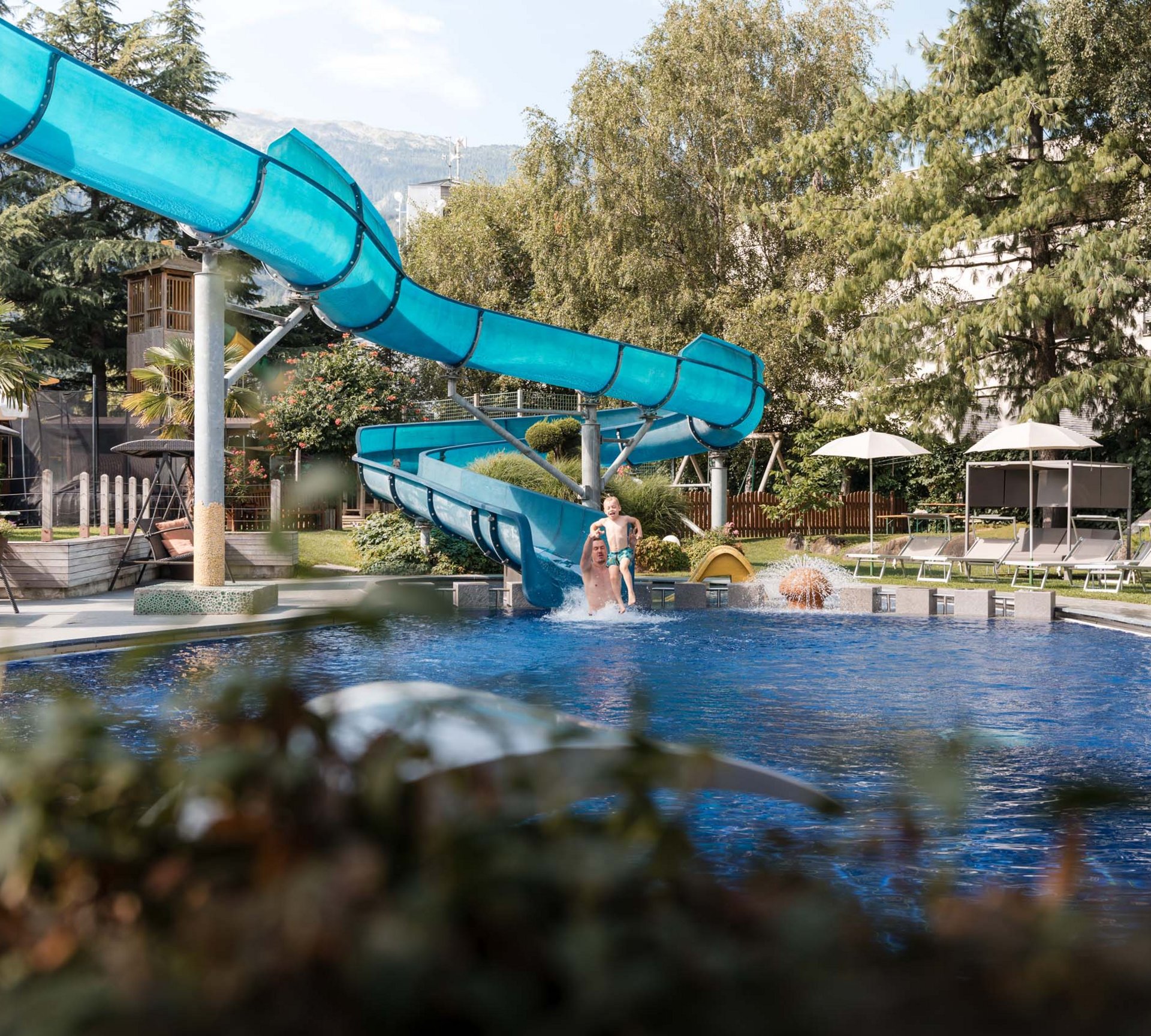 TYROL Family Retreat als Familienhotels in Südtirol Vater und Kind springen ins Schwimmbecken von Wasserrutsche im Garten