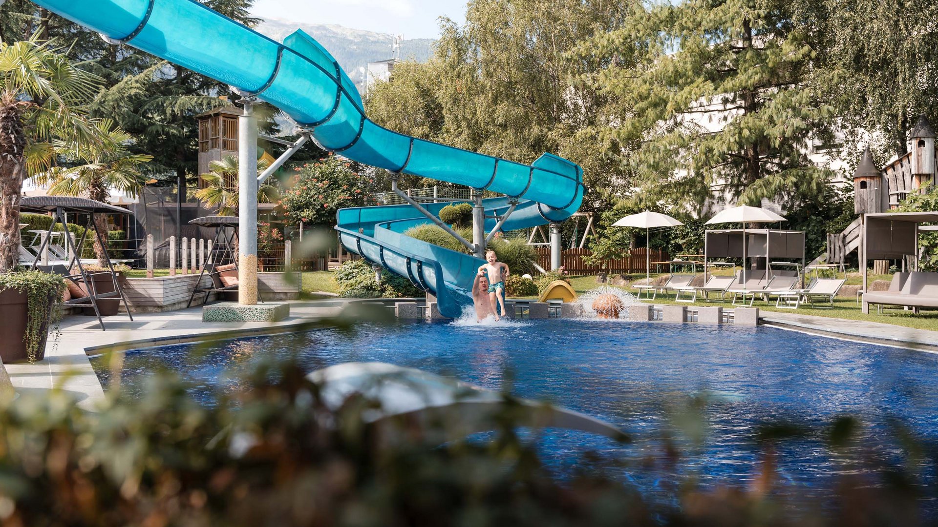 Ab ins Familienhotel in Südtirol mit Wasserrutsche! Vater und Kind springen ins Schwimmbecken von Wasserrutsche im Garten