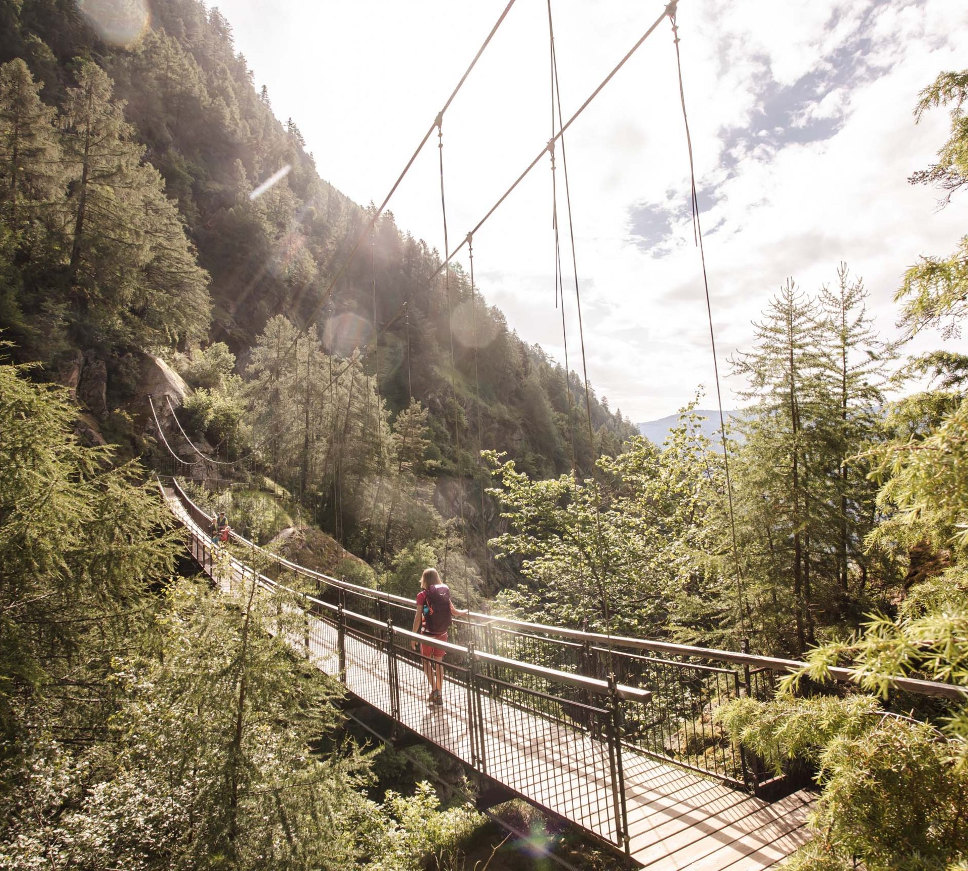 Gesunde Bewegung im Sporthotel in Naturns Person wandert über eine Hängebrücke in einem bewaldeten Gebirgstal bei Sonnenschein