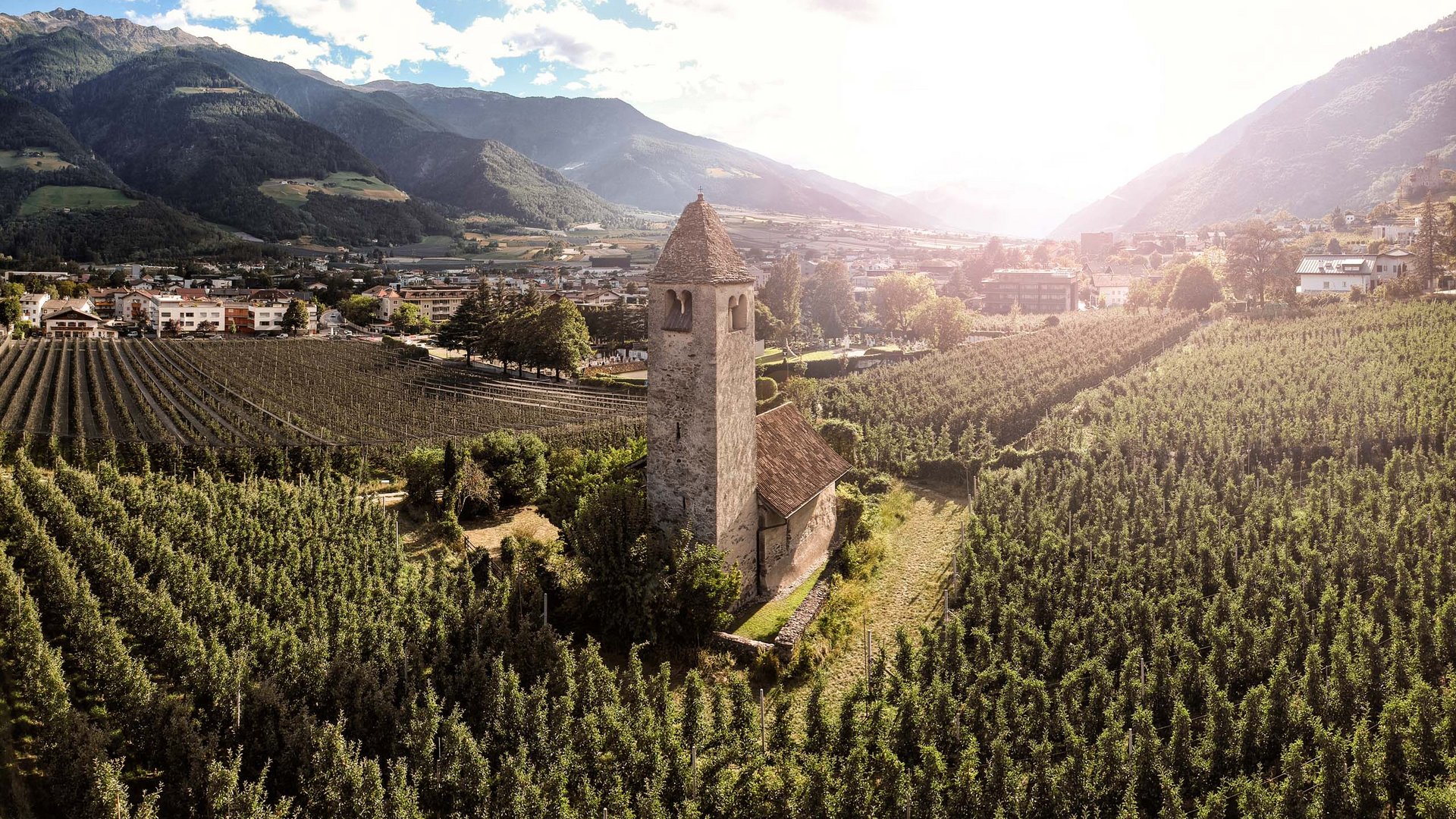 Euer vielseitiger Urlaub in Naturns Historischer Kirchturm inmitten von Apfelplantagen mit Bergen im Hintergrund
